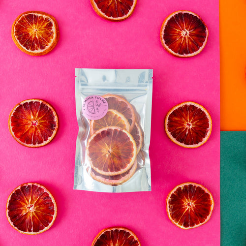 A small pack of dehydrated blood orange on a pink-orange-green background with slices of dehydrated oranges arranged around it.