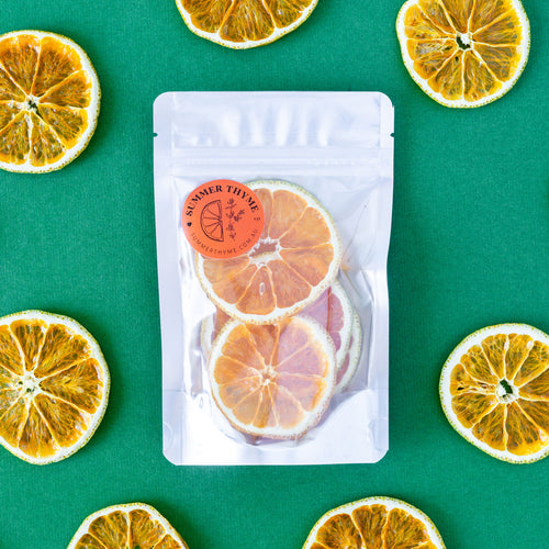 A small pack of dehydrated Valencia Oranges on a green background with dehydrated orange wheels arranged around it.