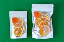 Load image into Gallery viewer, A small and a large pack of dehydrated Valencia Oranges arranged next to each other on a green background.
