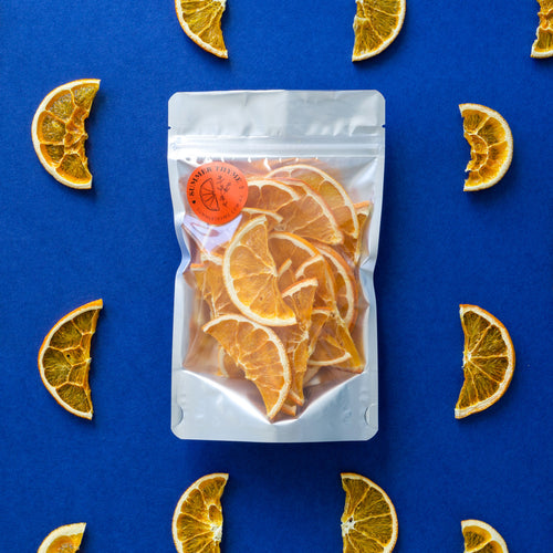 A large bag of dehydrated orange half-wheel slices on a dark blue background with half wheel dehydrated orange slices arranged around it.